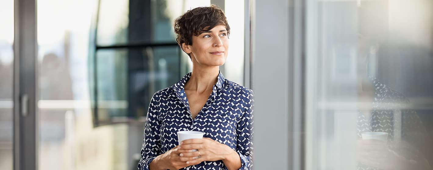 Eine Frau mit Kaffeetasse in der Hand lehnt sich an einem Fenster an und sieht hinaus.